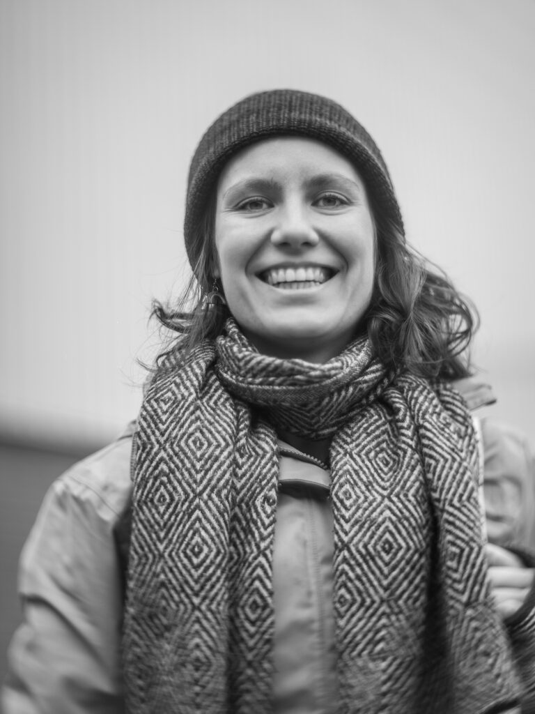 Schwarz-Weiß-Porträt der Illustratorin und Musikerin Anna Neuss. Sie lächelt in die Kamera und trägt eine dunkle Strickmütze, einen gemusterten Schal und eine helle Jacke.