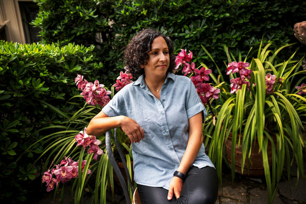 Hila Amit sitzt in hellem Oberteil und dunkler Hose, sanft lächelnd vor einer Grünpflanze mit rosa Blüten