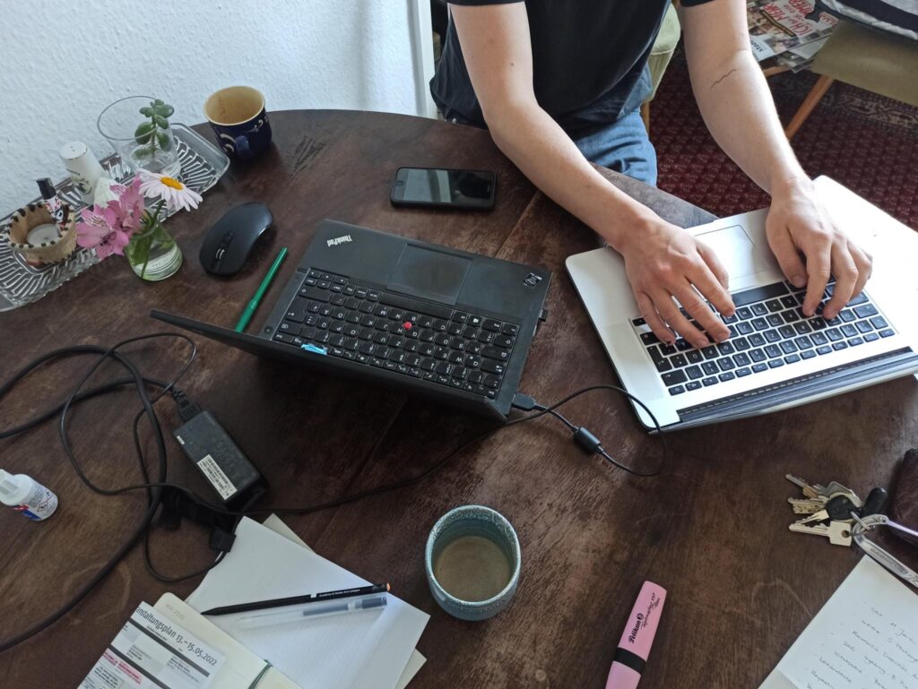 Das Foto zeigt eine Person, die an einem Runden Tisch am Laptop sitzt und arbeitet. Auf dem Tisch sind verschiedene gegenstände versträut: Stifte, Schlüssel, eine Teetasse, Zettel etc.