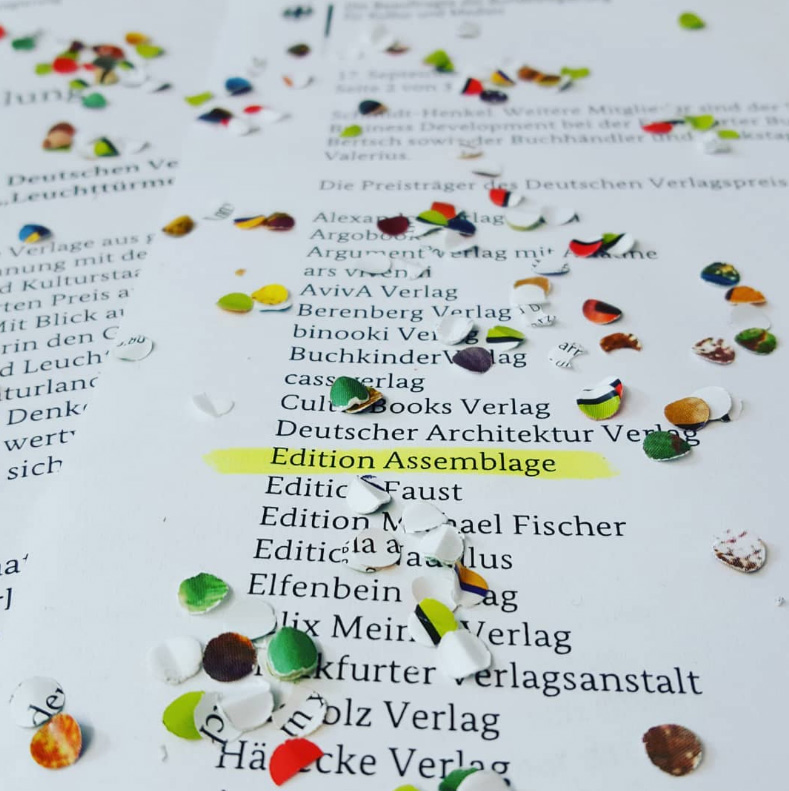 Auf einem weißen Blatt stehen in Computerschrift die Namen von 18 Verlagen. Edition Assemblage steht mittig und ist mit gelbem Texmarker markiert. Auf dem Zettel liegt verstreut buntes Konfetti.